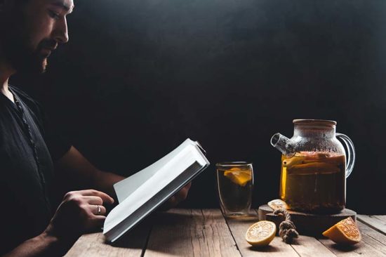Ein Mann liest ein Buch an einem Tisch. Vor ihm steht eine Teekanne mit Teeglas