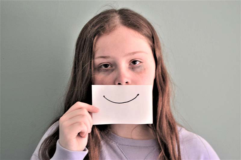 Thema Suizid: Ein Mädchen hält sich ein Papier mit einem aufgemaltem Lächeln vor das erschöpfte, leidende Gesicht