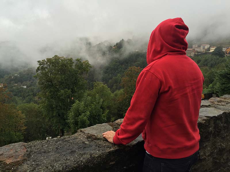 Leiden: Ein Mann mit rotem Kapuzenshirt steht mit dem Rücken im Bild an einer Mauer und blickt in ein grünes Tal im Nebel