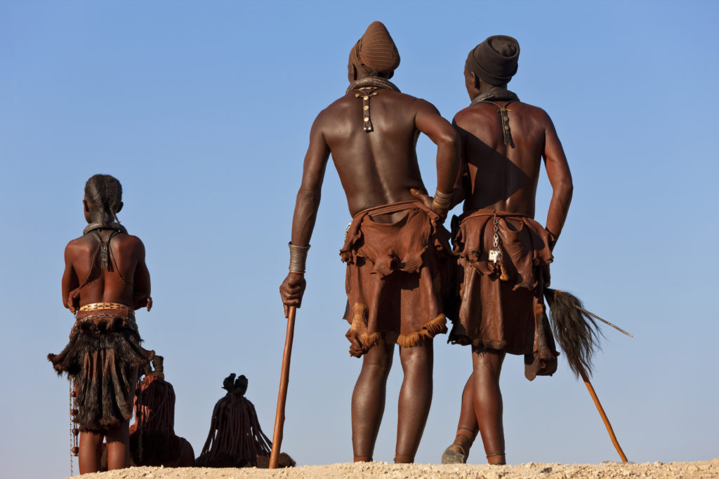 Kleine Gruppe von Himba-Männern, die traditionelle Kleidung tragen und in einer Wüste stehen.