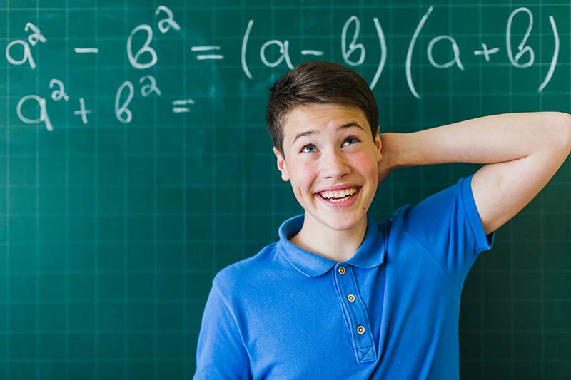 Ein junge kratzt sich am Kopf vor einer Tafel mit mathematischen Formeln. Thema: Hausaufgaben und wie man sie löst