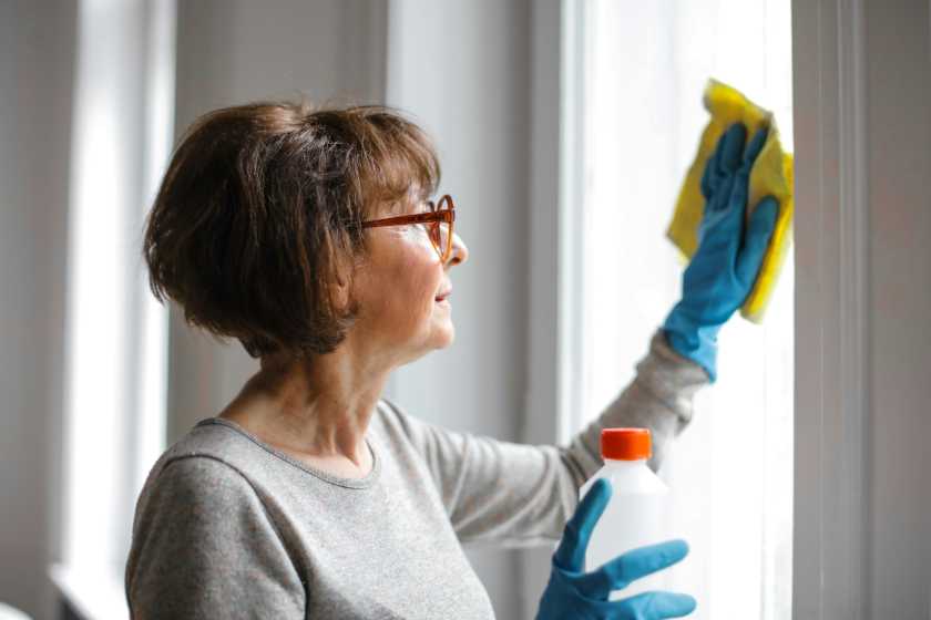 Eine reifere Frau trägt blaue Haushaltshandschue und putzt mit einem gelben Tuch ein Fenster 